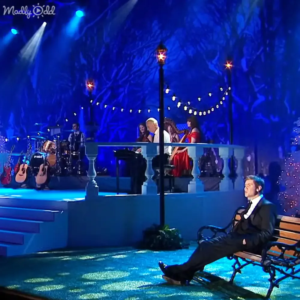 A man in a suit sits on a bench, gazing at a stage with musicians and a band under blue lighting.