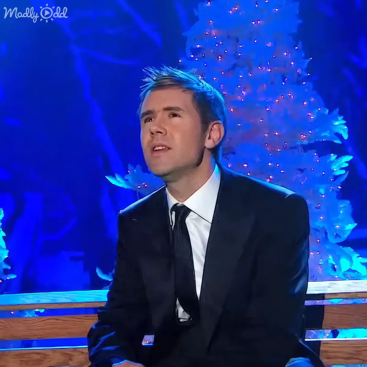 A man in a suit sits on a stage with a blue-lit background, possibly during a performance or event.
