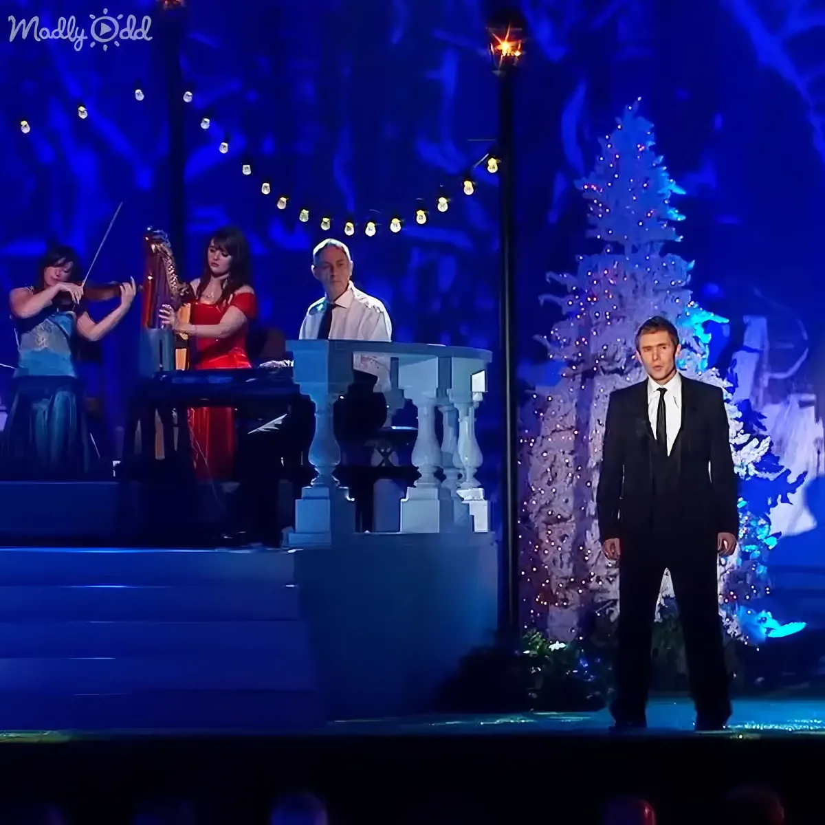 A man in a suit stands on stage, surrounded by musicians and a Christmas tree backdrop.