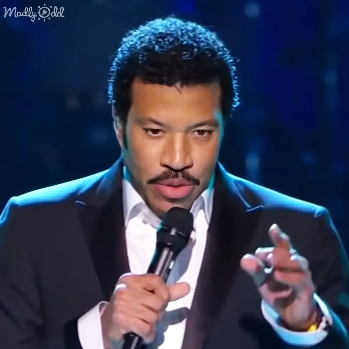 A man in a suit holds a microphone, gesturing with his right hand, against a dark background.