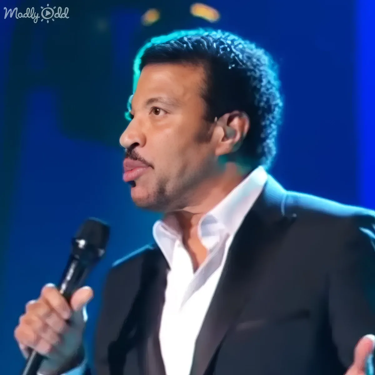 A man in a suit sings into a microphone on stage, illuminated by blue lights.