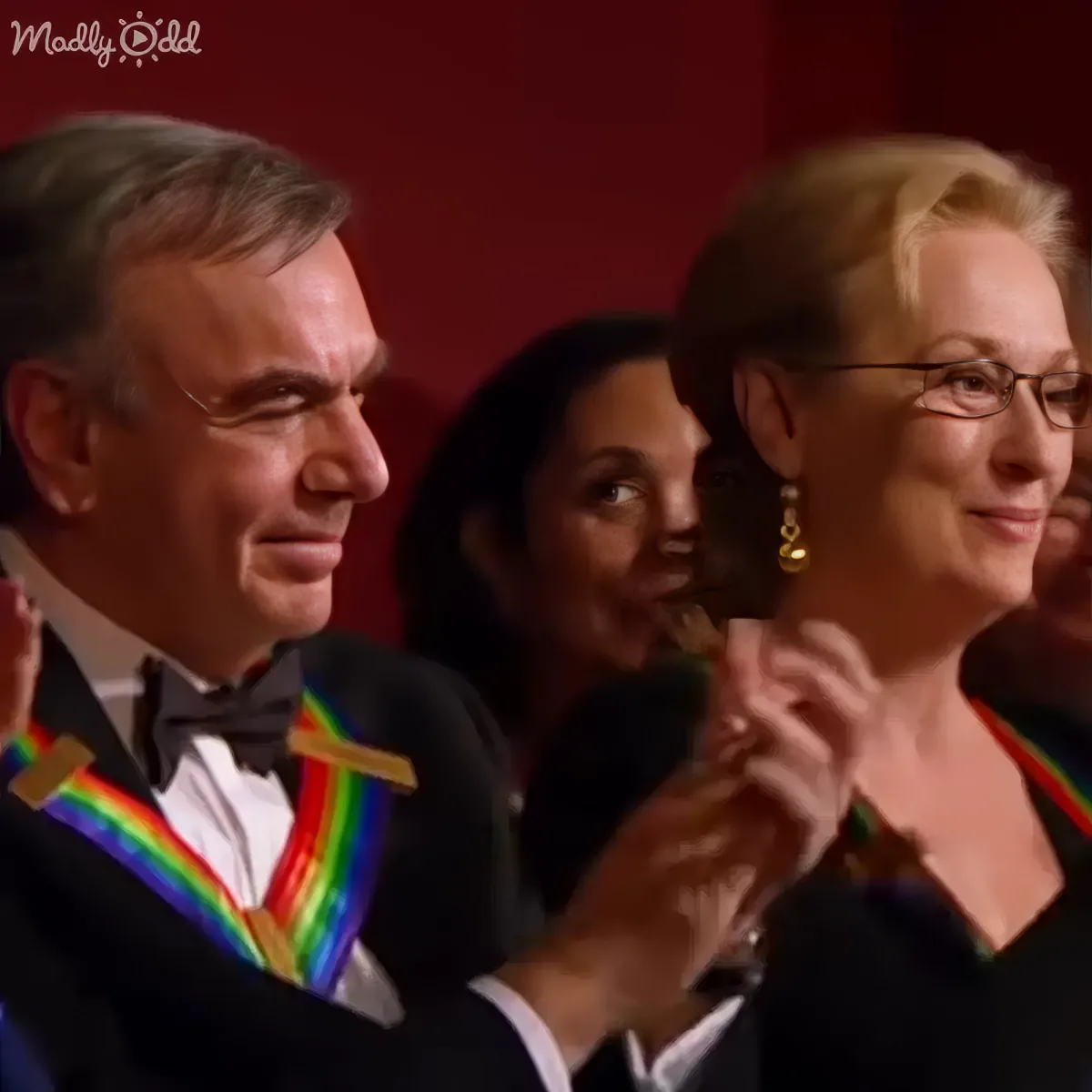 A man in a tuxedo and a woman in a black dress with rainbow ribbons around their necks are clapping.