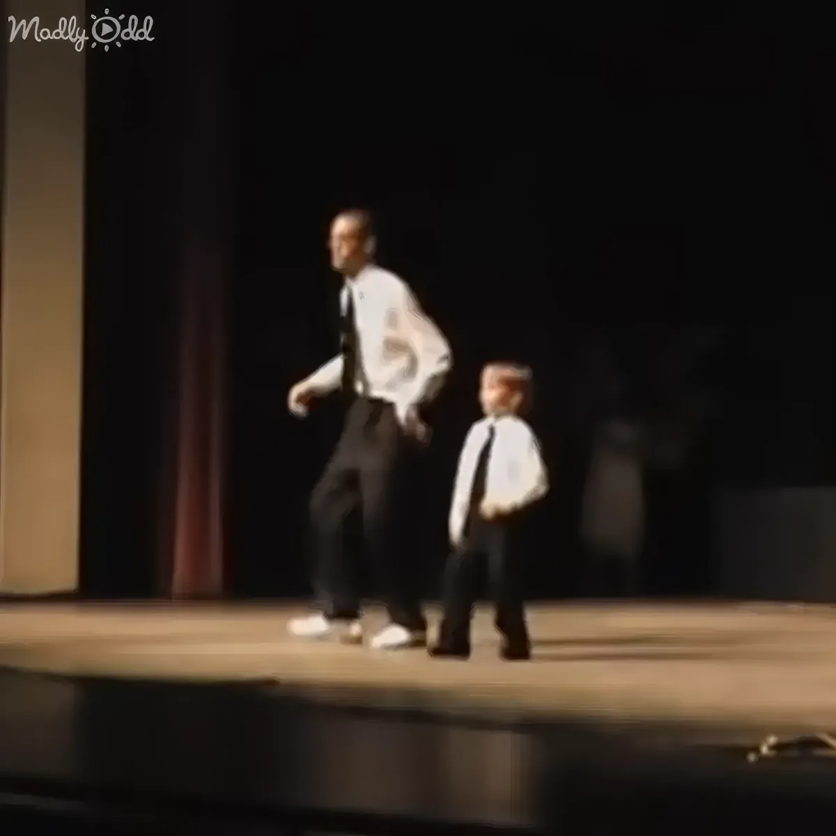 A man and a young boy in formal attire walk side by side on a stage.