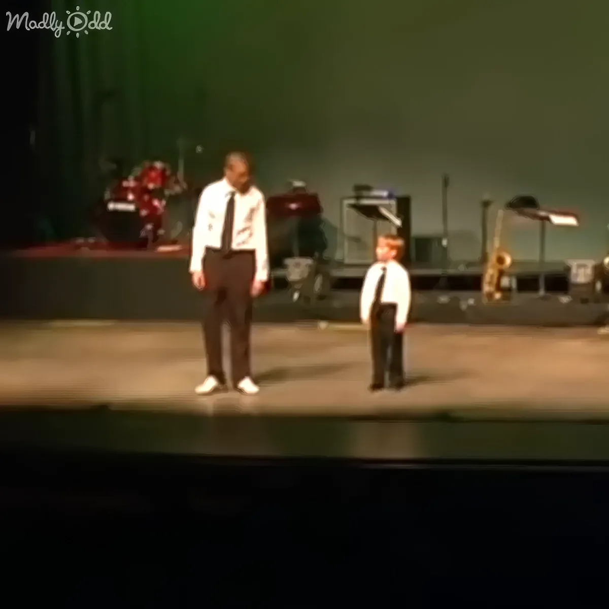 A man and a young boy in suits stand on stage, facing each other, with musical instruments in the background.