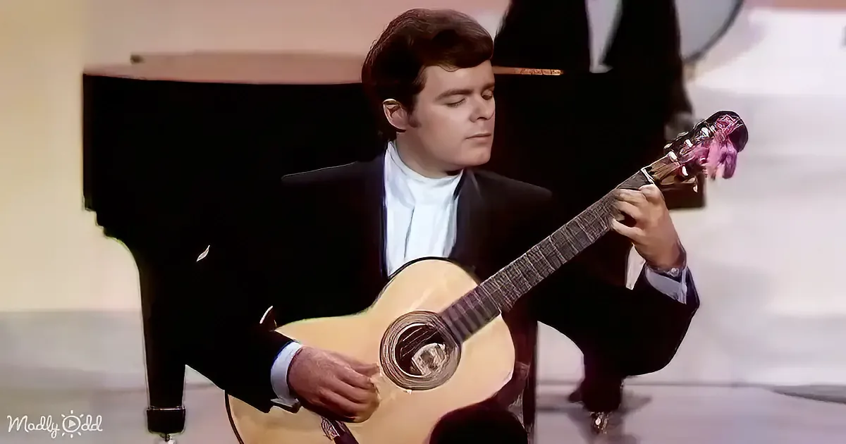 A man in a suit plays a classical guitar, his eyes closed, in front of a piano.