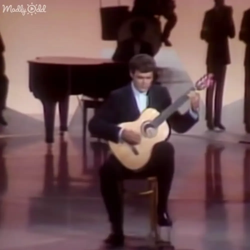 A man in a suit plays a classical guitar on a stool, surrounded by silhouettes of people and a piano.