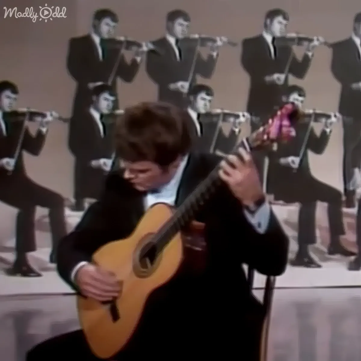 A man in a suit plays a guitar while a group of identical men in suits play flutes behind him.