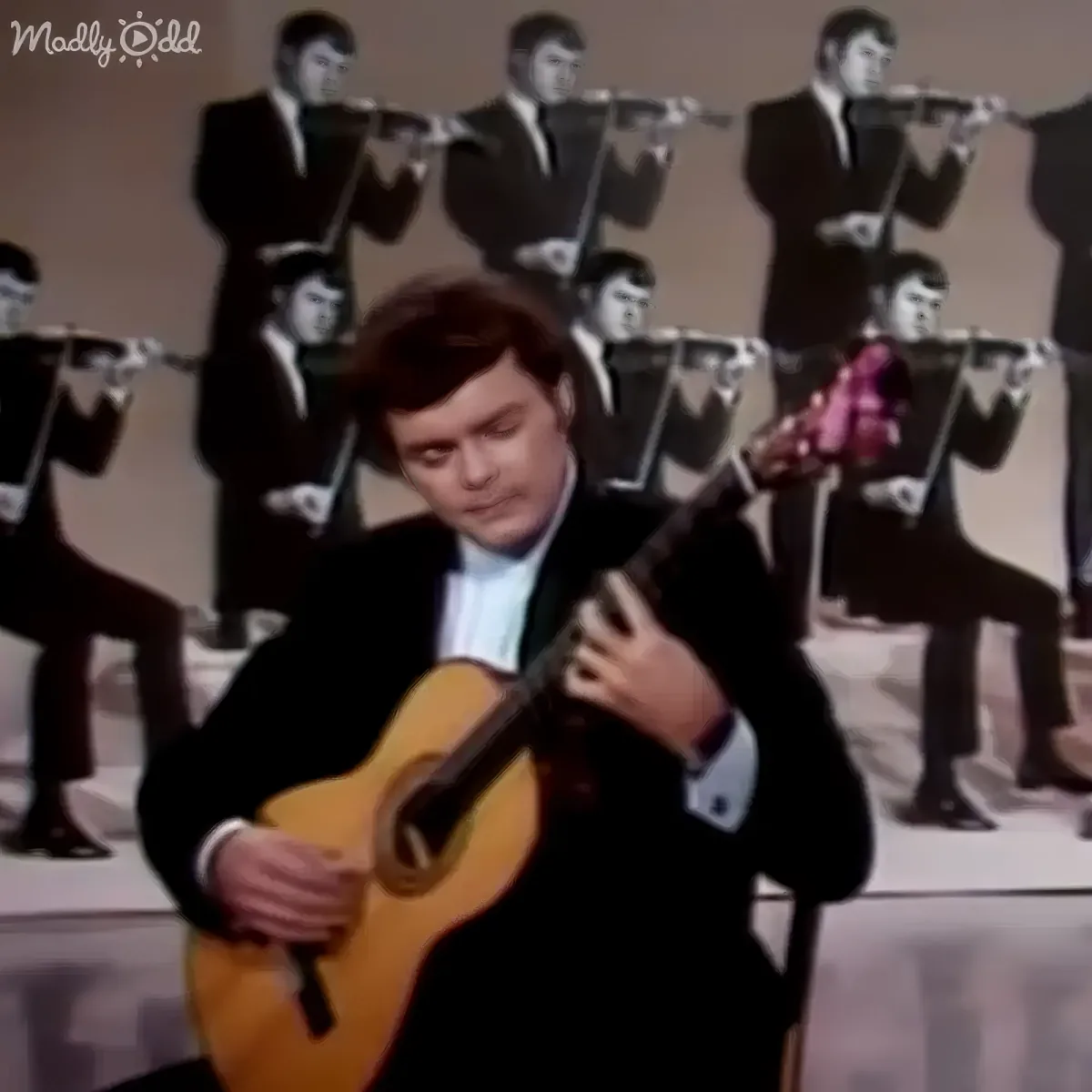 A man in a suit plays a guitar in front of a backdrop of identical flute players.