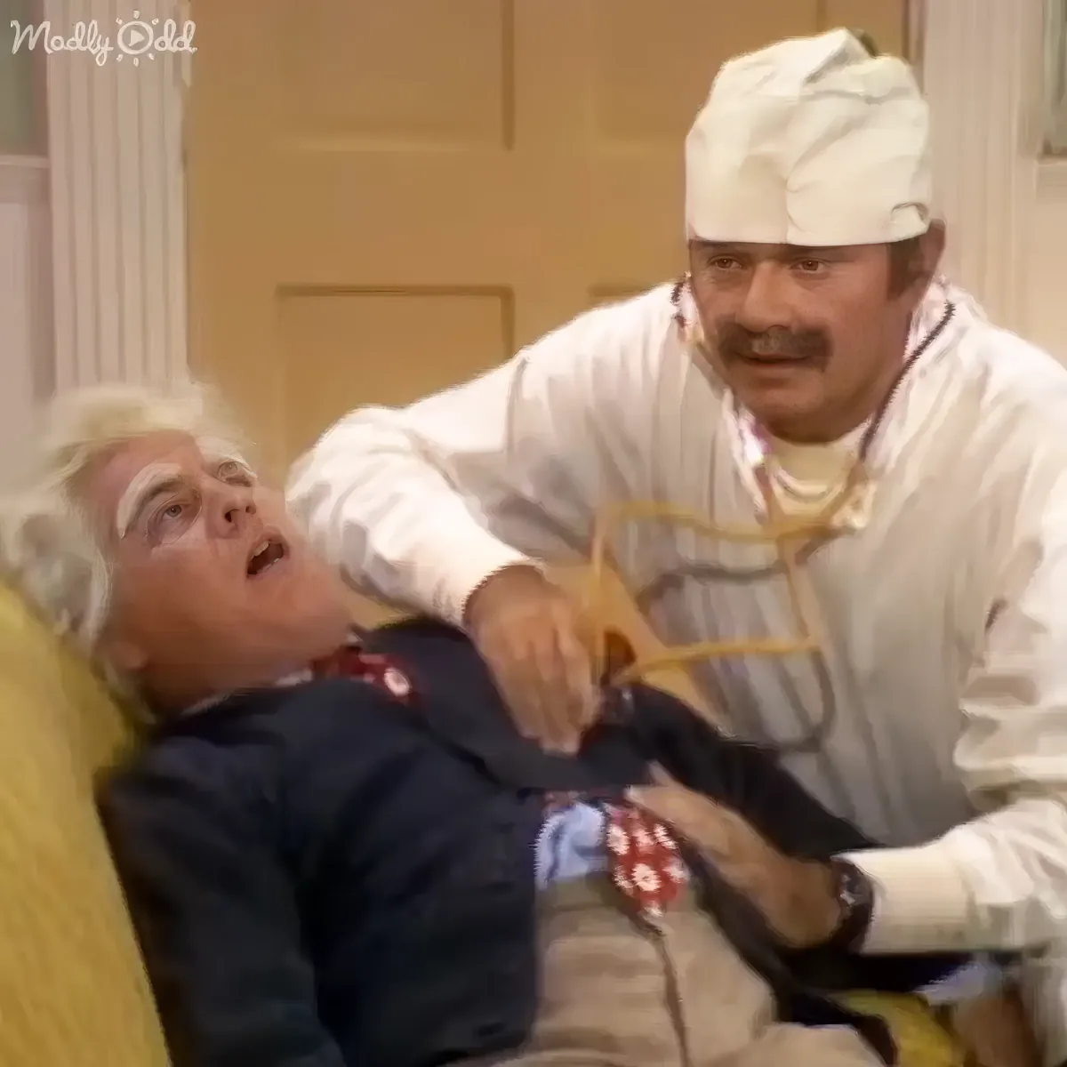 A man in a white chef's outfit is holding a spoon to another man's mouth, who is reclining on a yellow couch.