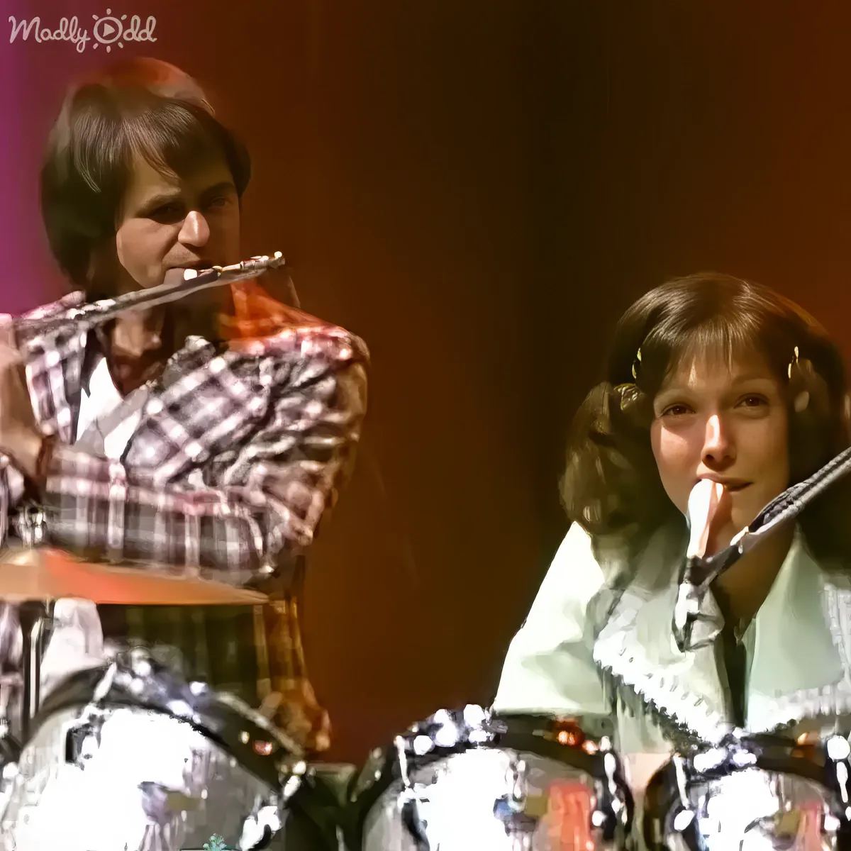 A person in a plaid shirt plays a flute, while another person in a white shirt plays a drum set.