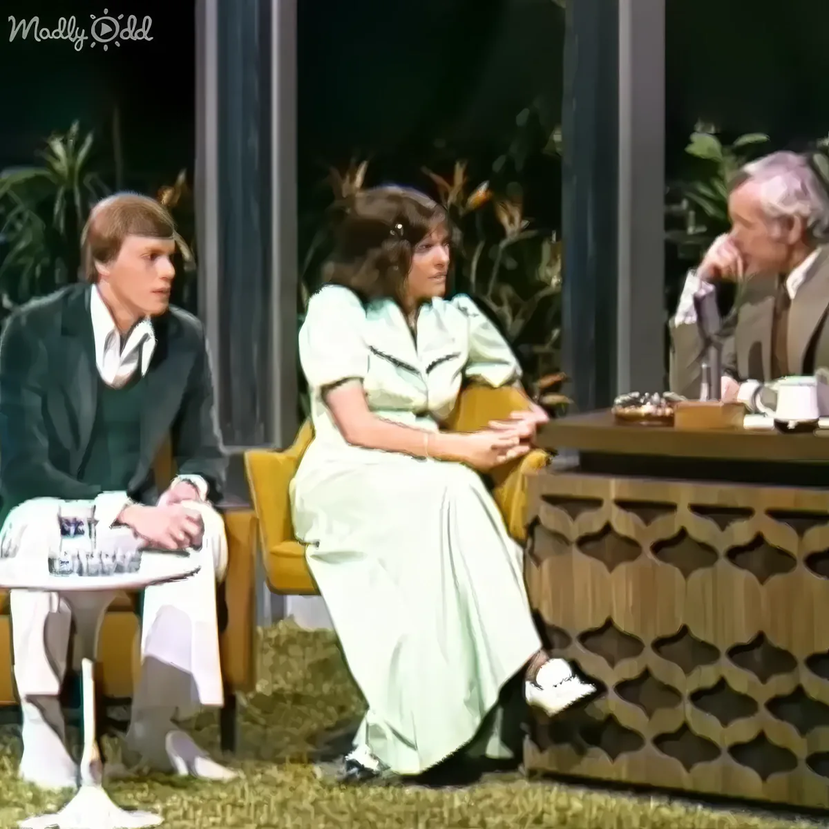 A woman in a light green dress sits on a yellow chair, talking to a man in a suit on a talk show set.