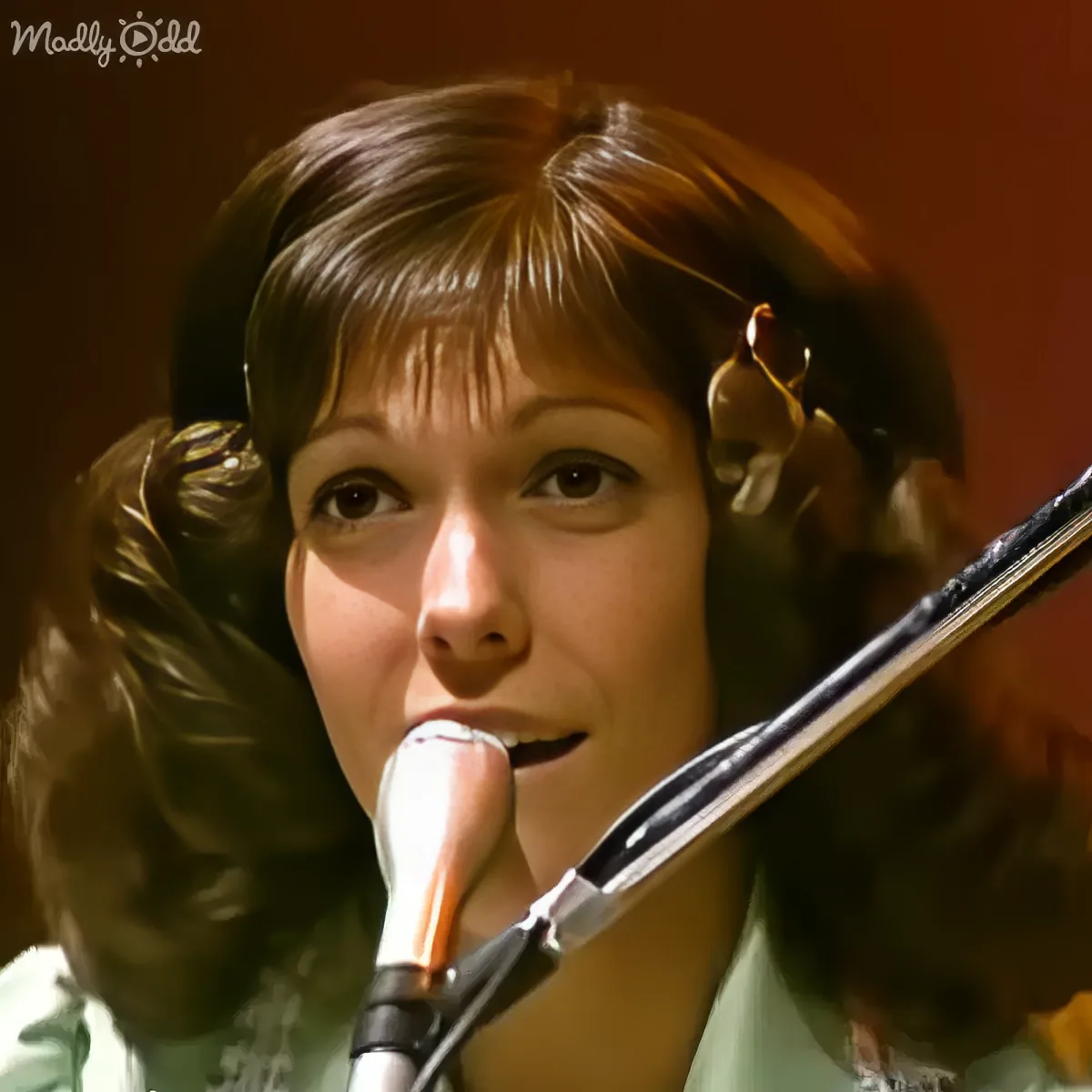 A woman with long, curly brown hair sings into a microphone, wearing a green top.
