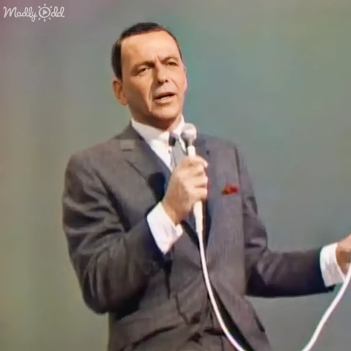 A man in a suit holds a microphone, gesturing with his right hand, against a plain background.