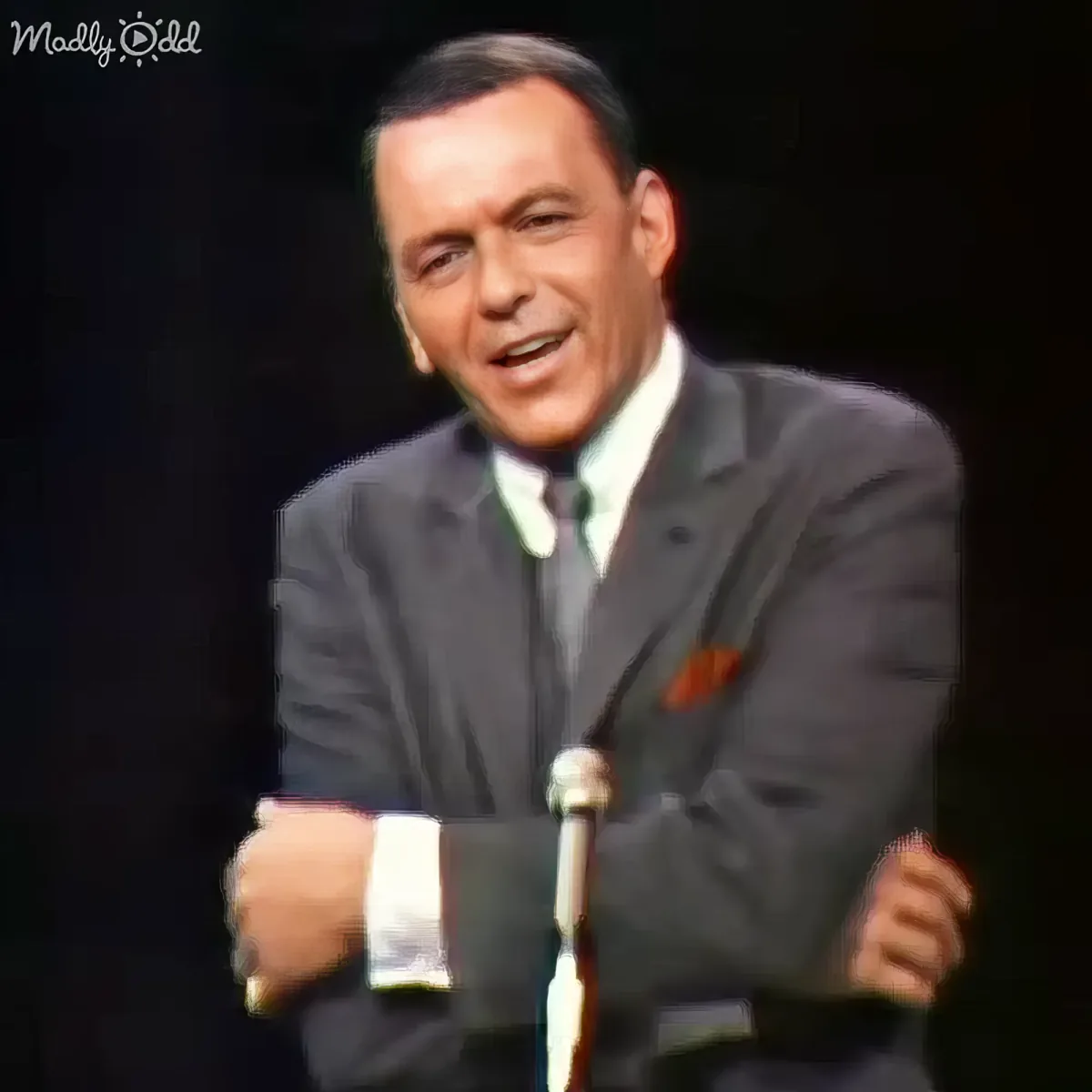 A man in a suit and tie is seated, smiling, with a microphone in front of him.