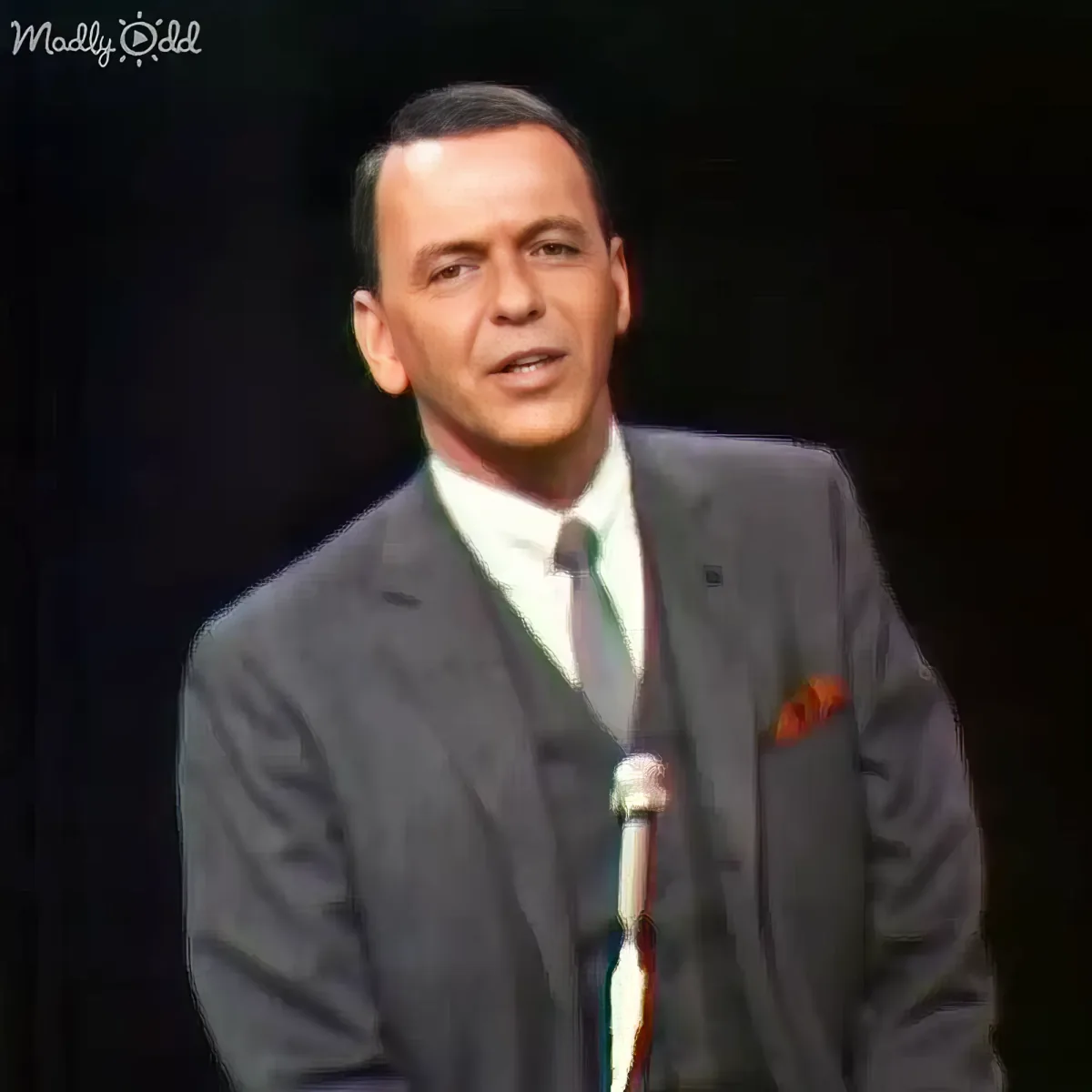 A man in a suit and tie stands on stage, speaking into a microphone.