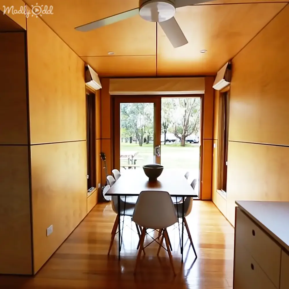 A modern dining area with a long table, white chairs, and a large sliding door leading to a green outdoor space.