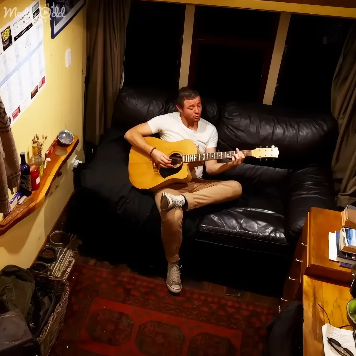 A man sits on a black leather couch playing an acoustic guitar in a cozy room with a red patterned rug.