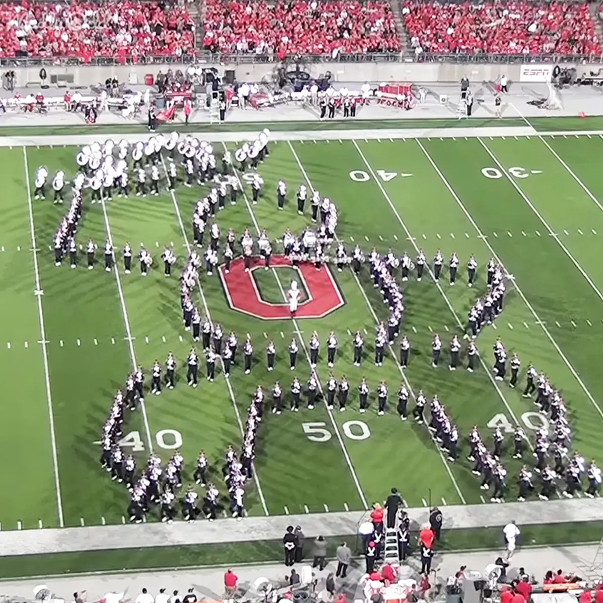 A marching band forms a large "U" on a football field, with spectators in the stands.