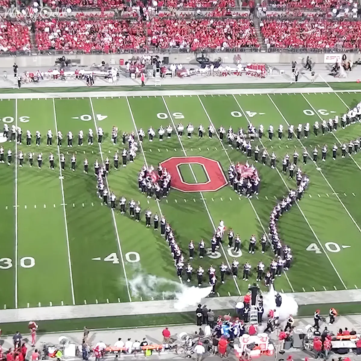 A marching band forms a large "O" on a football field with smoke effects.