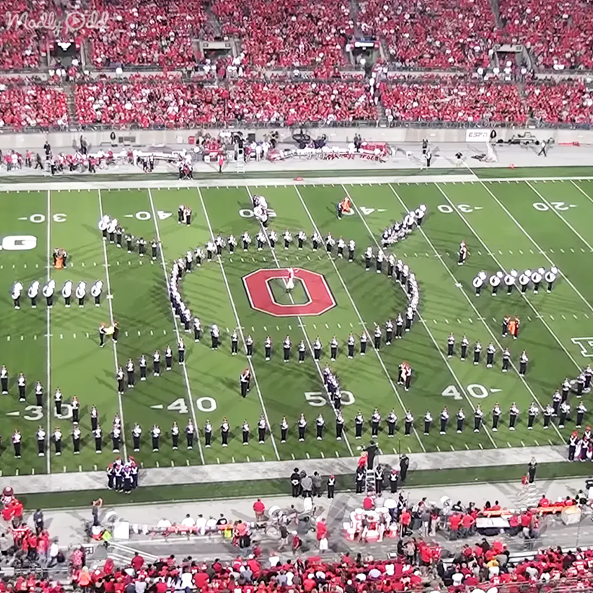 A marching band forms a large "O" on a football field, surrounded by a crowd of spectators.