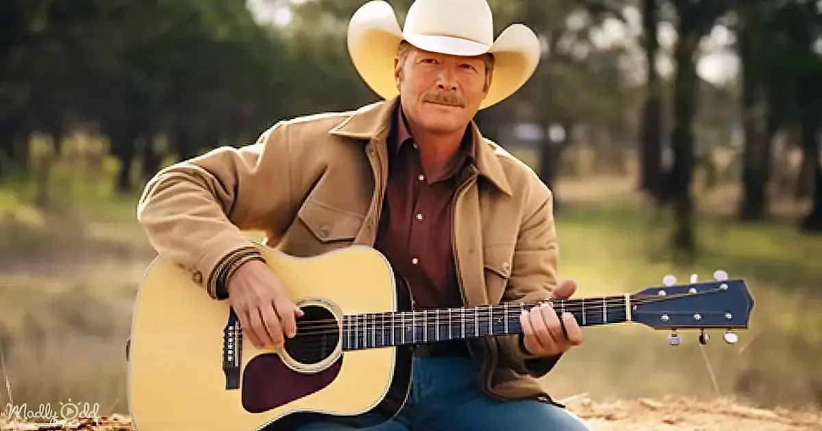 A man in a cowboy hat and tan jacket plays an acoustic guitar outdoors.