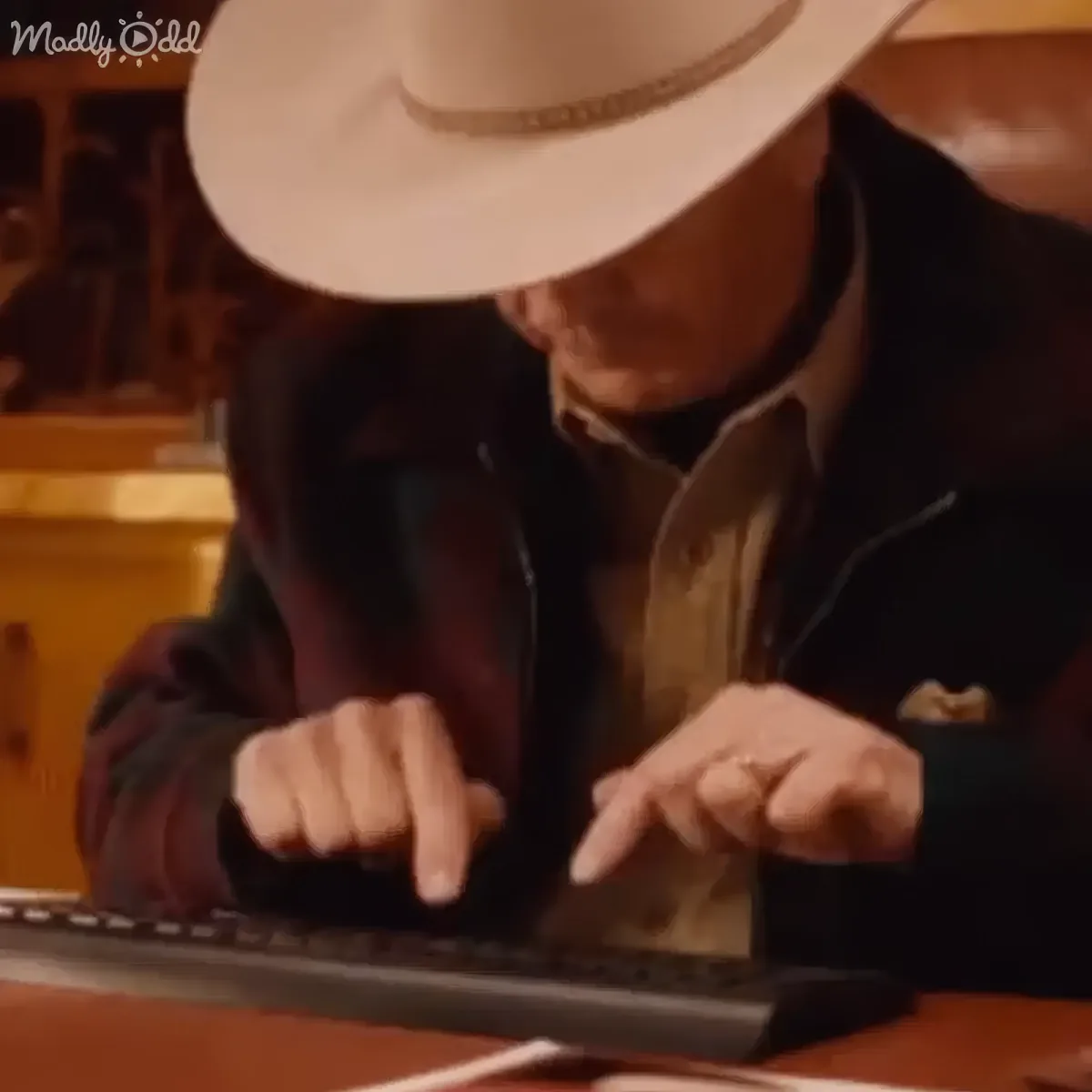An older man in a white cowboy hat types on a keyboard, wearing a dark jacket and a ring on his left hand.