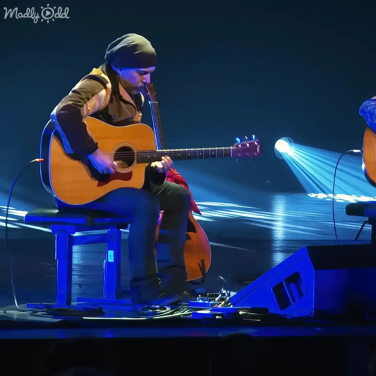 A musician plays an acoustic guitar on stage under blue lighting, surrounded by equipment.