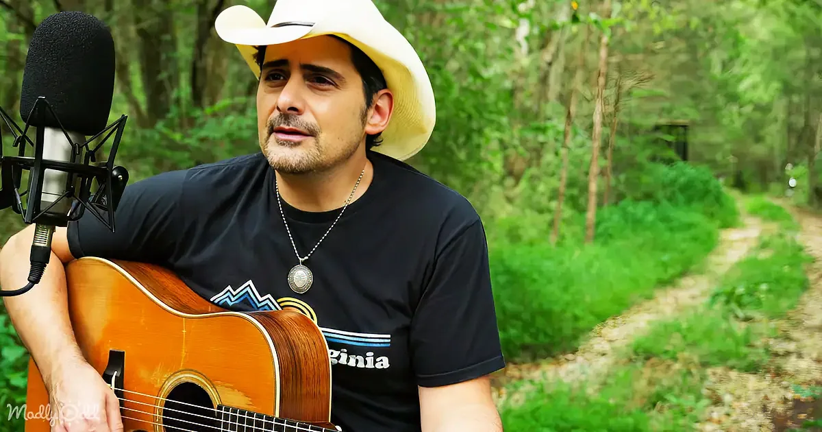 A man in a cowboy hat plays an acoustic guitar in front of a forest backdrop.