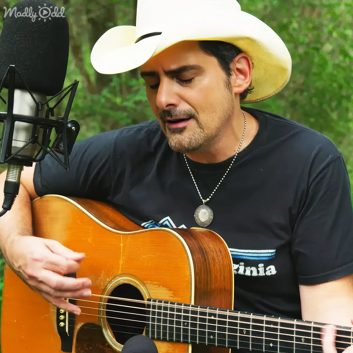 A man in a cowboy hat plays an acoustic guitar, singing into a microphone outdoors.