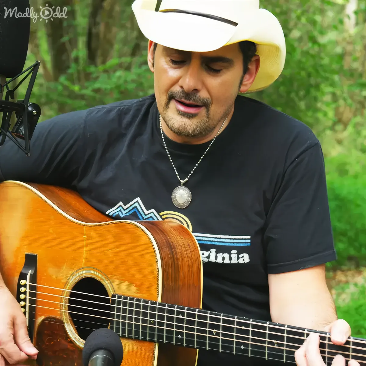 A man in a white cowboy hat plays an acoustic guitar, wearing a black "Virginia" t-shirt, with a microphone in front of him.