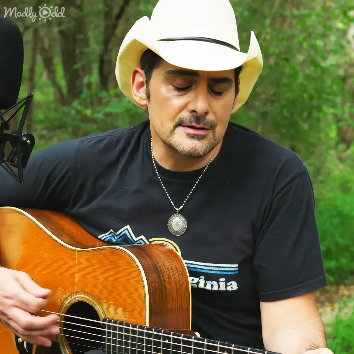 A man in a white cowboy hat and black shirt plays an acoustic guitar outdoors.