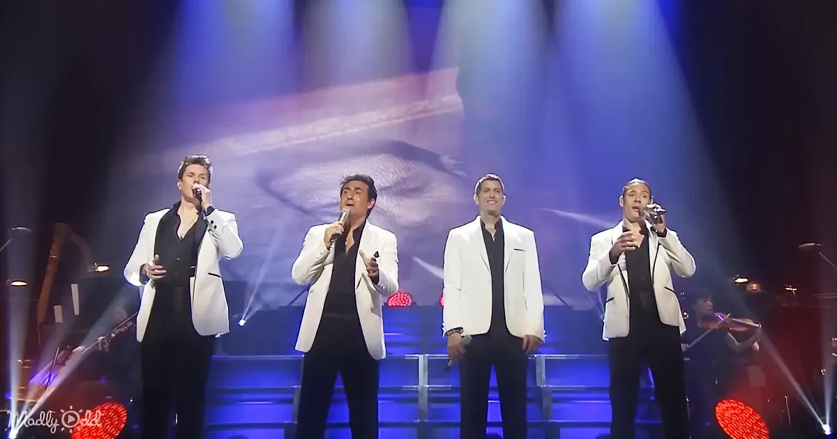 Four men in white jackets sing on stage with microphones, accompanied by a harp and violin.