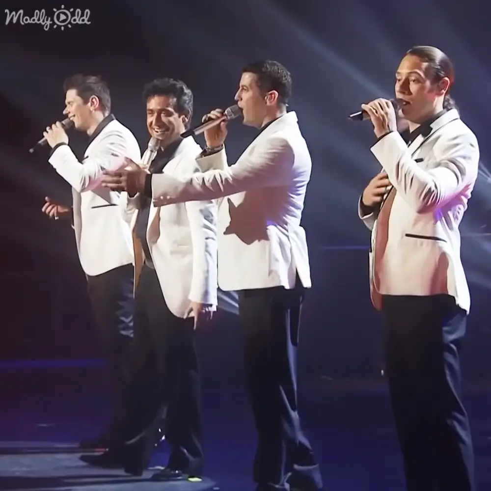 Four men in white jackets and black pants sing on stage with microphones, illuminated by stage lights.