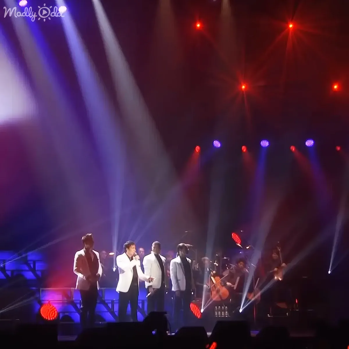 A group of performers in white suits sing on stage under vibrant red and blue spotlights.