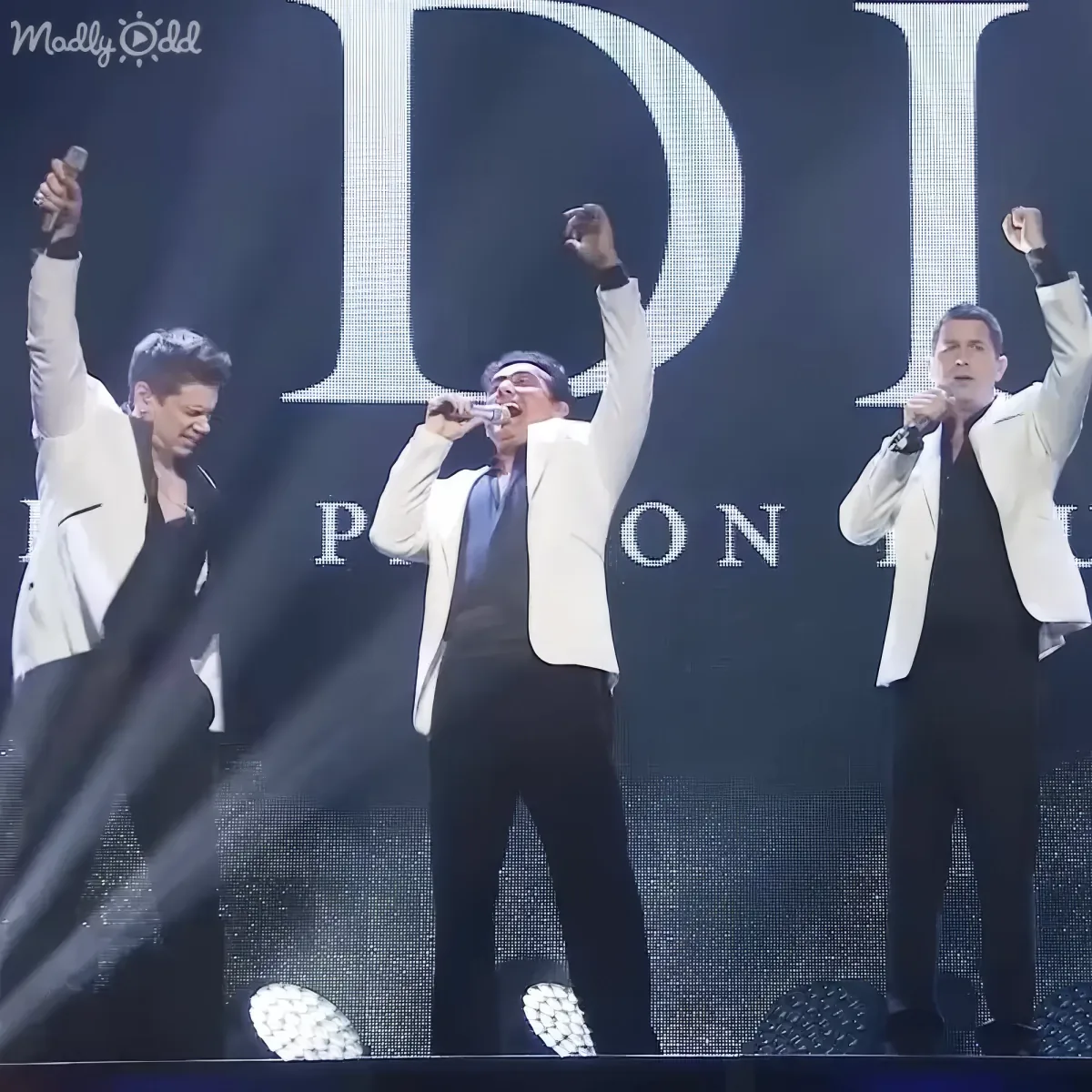 Three men in white jackets perform on stage with microphones, raising their fists in the air.