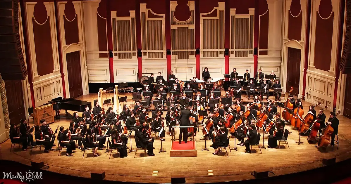 Conductor leads an orchestra in a grand hall with red and white decor.