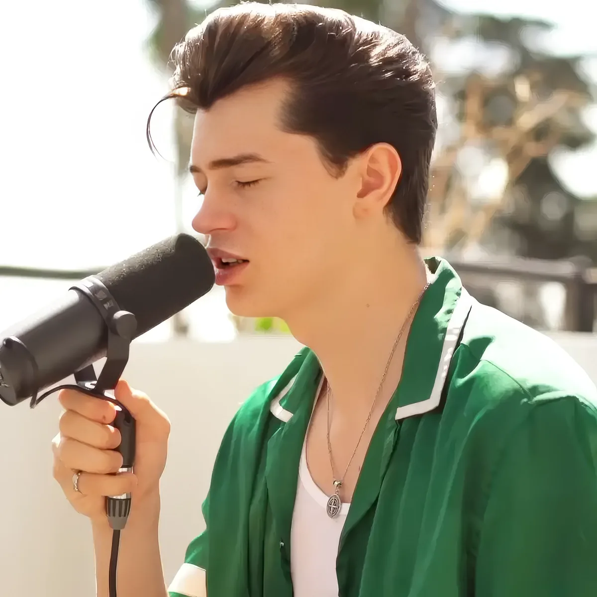 A person in a green jacket sings into a microphone, wearing a white shirt and a necklace.