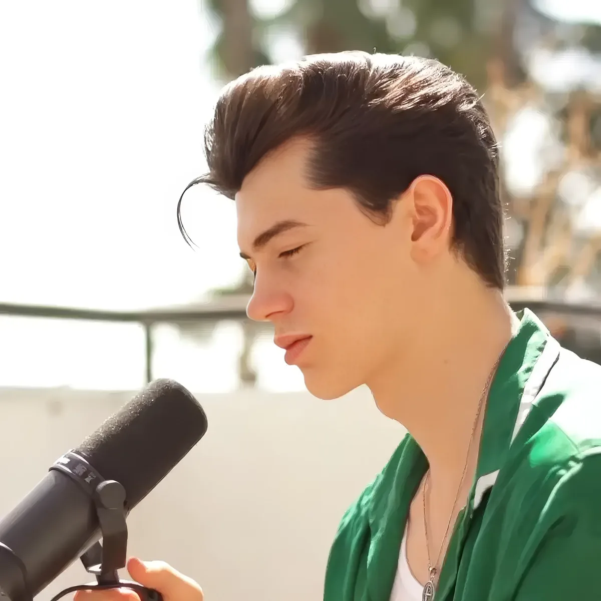 A person in a green shirt holds a microphone, looking down, with a bright background.