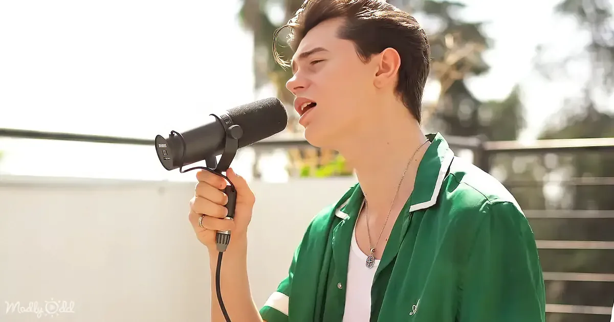 A person in a green shirt sings into a microphone outdoors, with trees and sunlight in the background.