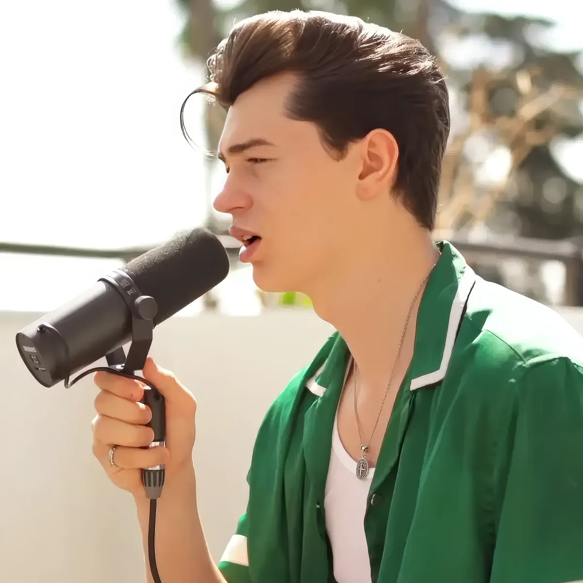 A person in a green shirt sings into a microphone outdoors, with a bright background.