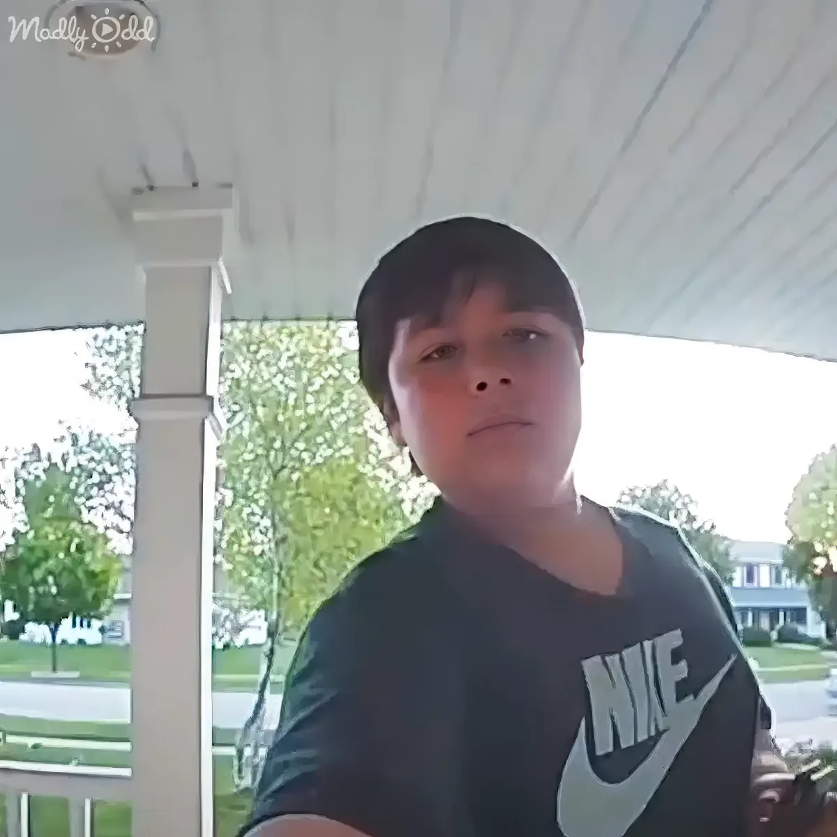 A boy in a Nike shirt sits on a porch, looking directly at the camera with trees and a house in the background.
