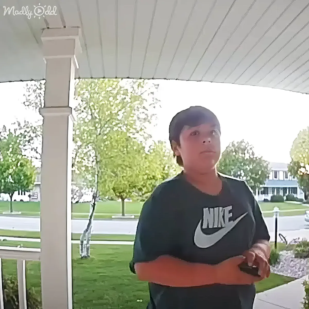A person in a Nike shirt stands on a porch, holding a phone, with trees and a house in the background.