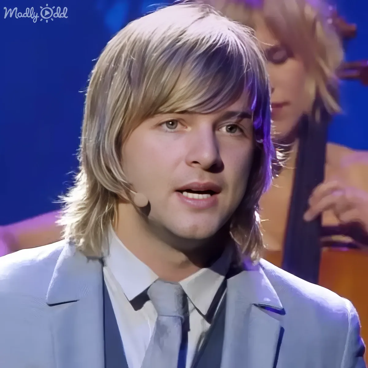 A man with long blonde hair and a light blue suit is speaking on stage, with a musician in the background.