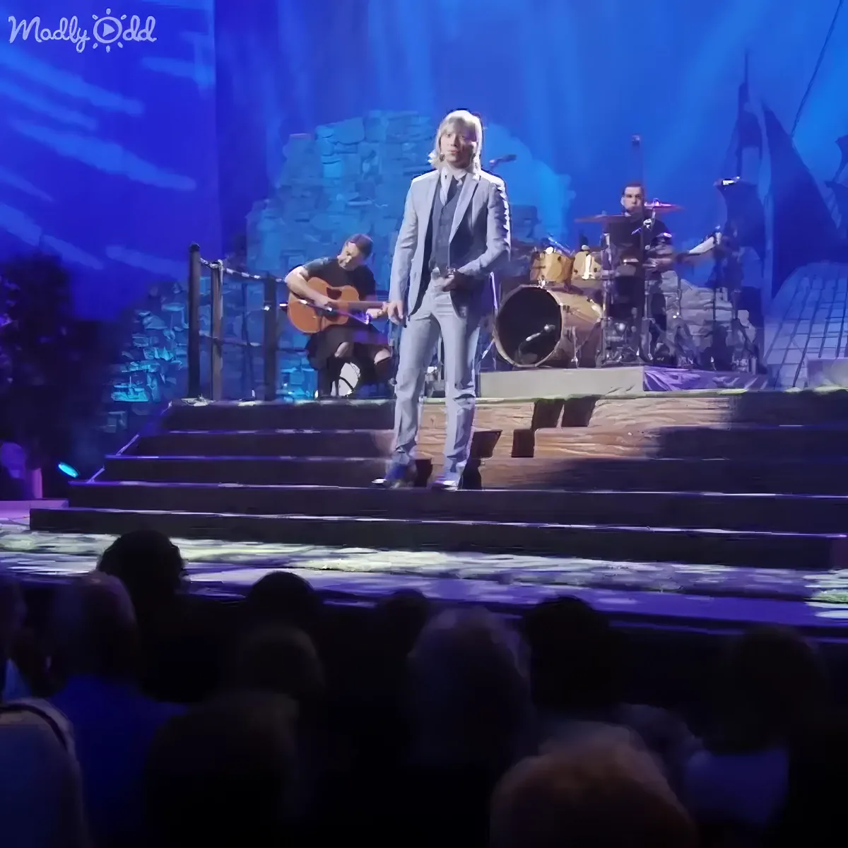 A man in a suit stands on stage, surrounded by musicians and a scenic backdrop.