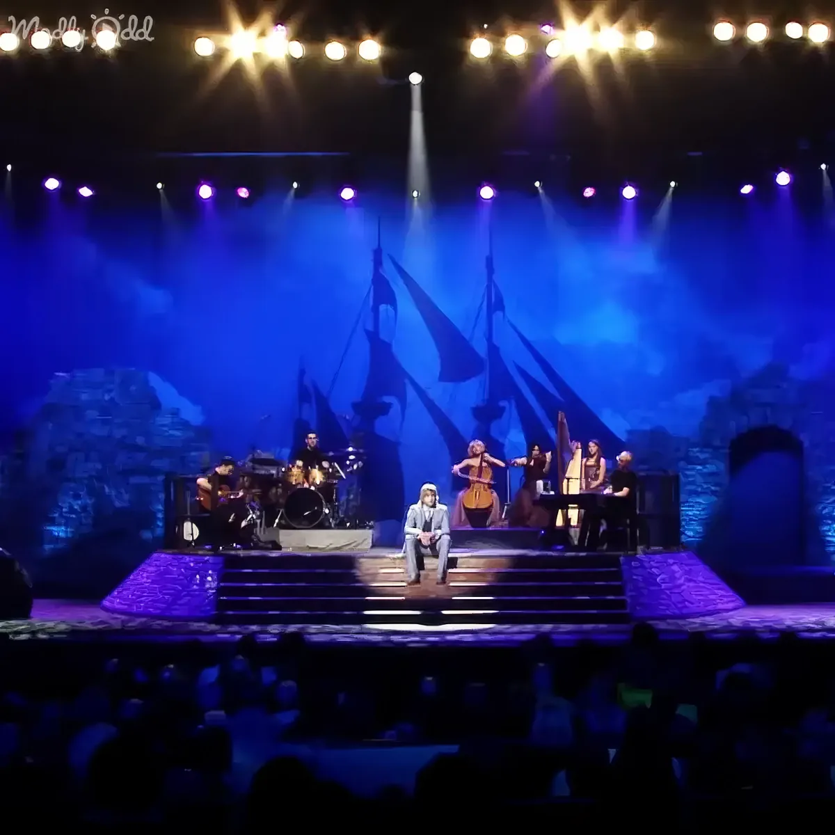 A performer sits center stage with musicians behind, illuminated by blue and white lights, against a backdrop of a ship...