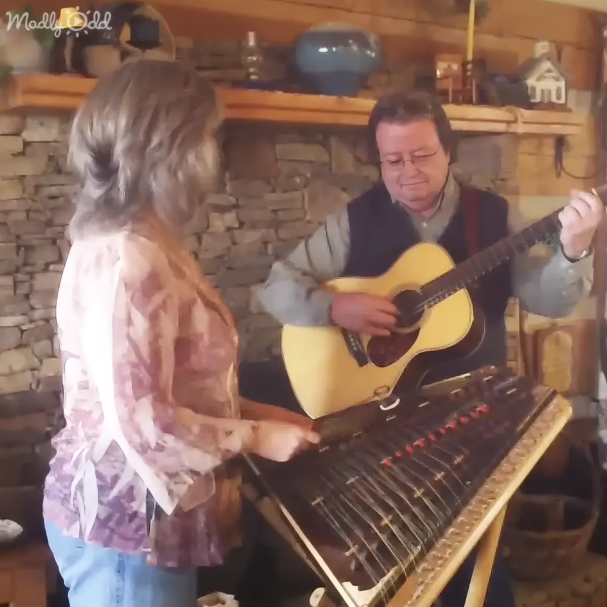 A man plays guitar while a woman plays a stringed instrument in a cozy room.