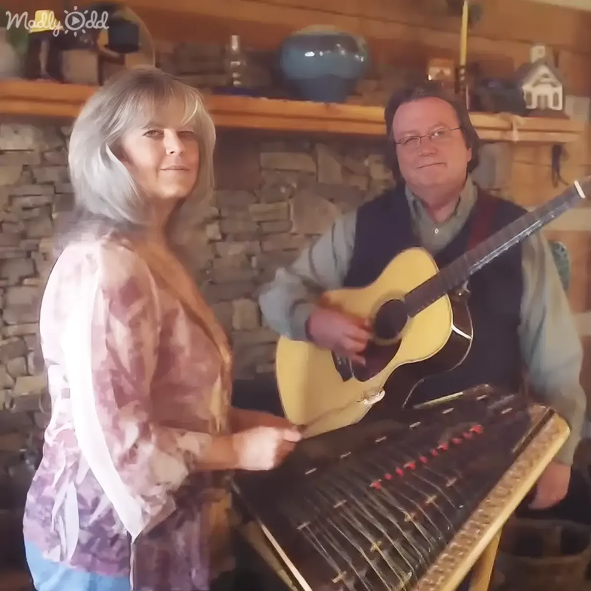 A woman plays a hammered dulcimer while a man holds a guitar in a cozy room with stone walls.