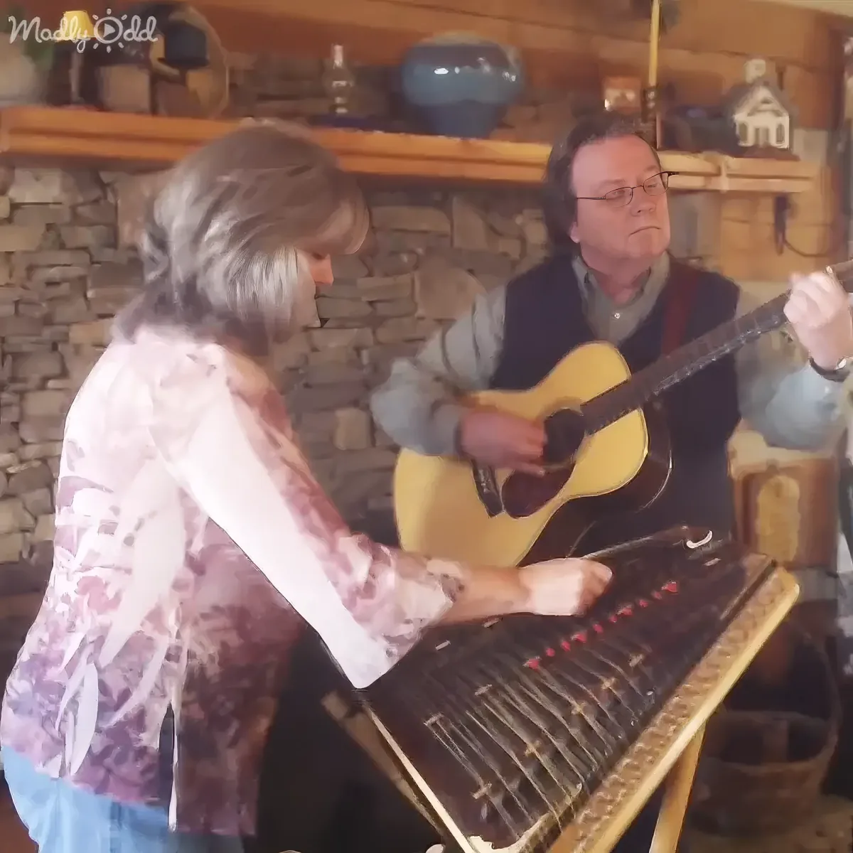 A woman plays a keyboard while a man strums a guitar in a cozy, rustic setting.