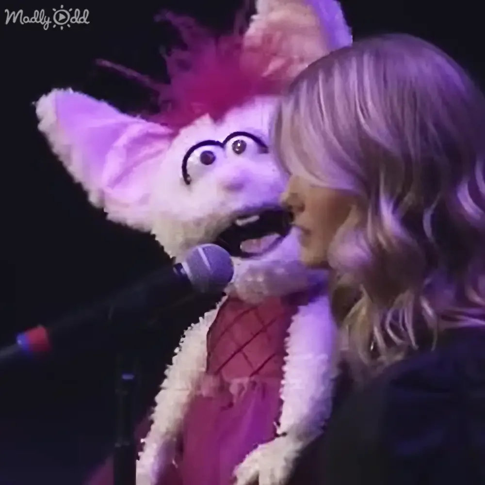 A puppet with large ears and a red dress sings into a microphone, accompanied by a person with long hair.