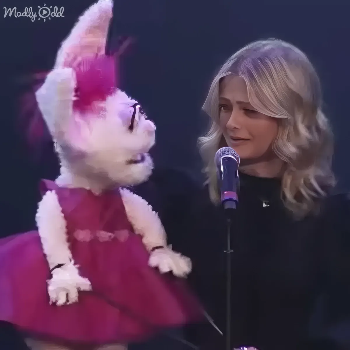 A puppet in a pink dress and a woman with blonde hair stand near a microphone on stage.